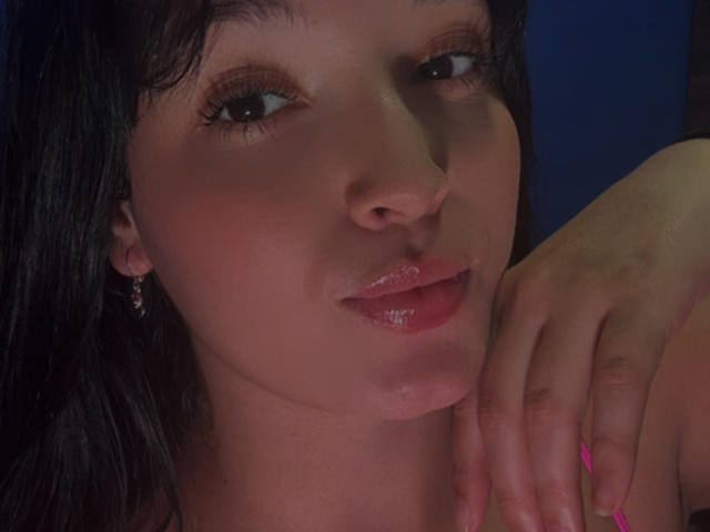 SarahGood's face with shiny pink lips, dark eyes, and glossy black hair. She's touching her cheek with pink-manicured fingers, wearing small earrings. Her skin is slightly flushed.