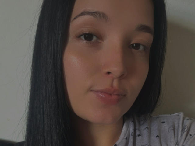 SarahGood has medium brown eyes, black straight hair, and a slight smile with pinkish lips. She's wearing a grey shirt with small black patterns. Her skin is light.