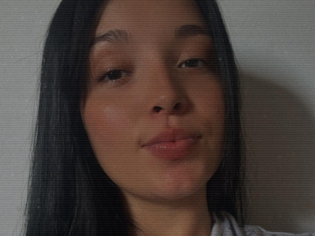 SarahGood's face with black hair, light brown skin, and a subtle smile, showing her slightly parted lips and straight eyebrows, in a blurry, grainy photo.
