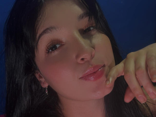 SarahGood with glossy pink lips, dark wet-looking hair, and a hand near her mouth, smiling slightly. She has a cute face, small nose, and light skin.