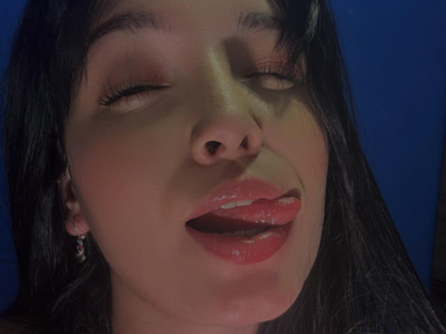 SarahGood's face with eyes closed, shiny pink lips slightly open, tongue touching upper lip, dark straight hair, small hoop earrings, blue background, slight shadow on her nose.