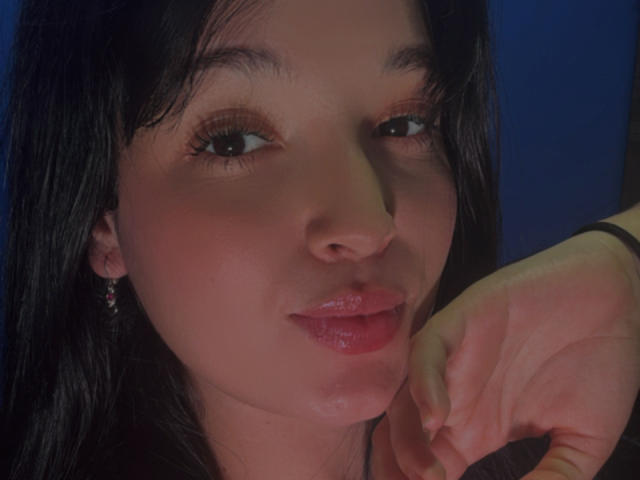 SarahGood has shiny pink lips, dark wet-looking hair, and is touching her chin with a slightly flushed face, brown eyes, and small pink earrings against a blue background.