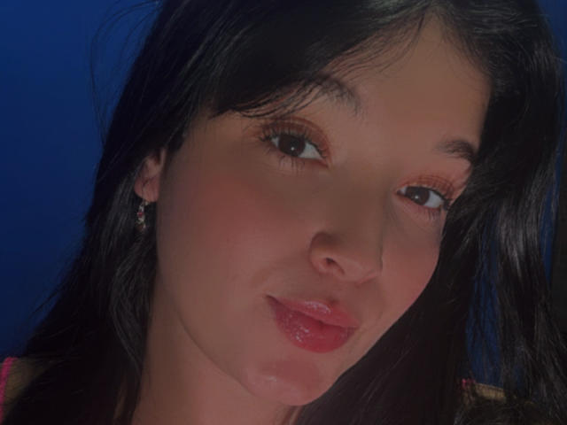SarahGood has dark hair, brown eyes, and pinkish-red lips. She's wearing small earrings and has a slight smile with a blue background. Her nose is slightly shiny.