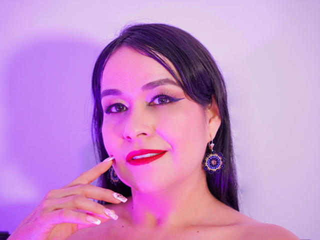 TamaraFoxy has black hair, red lipstick, and is smiling with her finger near her mouth. She's wearing purple earrings and has pink-purple lighting on her face.