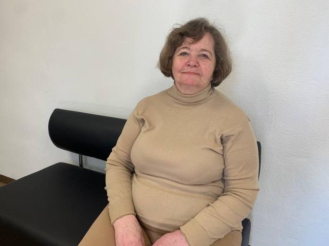 ZoryanaPtah, een oudere vrouw met kort bruin haar, zit op een zwarte stoel, gekleed in een beige coltrui die haar kleine borsten en een licht mollig achterwerk laat zien..