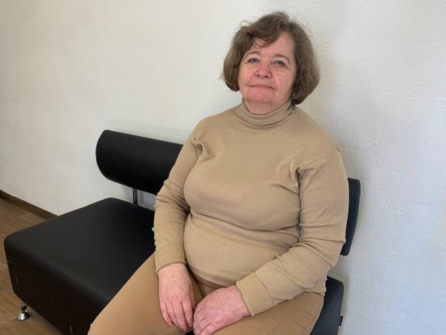 ZoryanaPtah heeft kort bruin haar, een beige trui en beige broek, en zit op een zwarte stoel met een neutrale uitdrukking, waarbij een licht mollig, middelbaar postuur zichtbaar is..