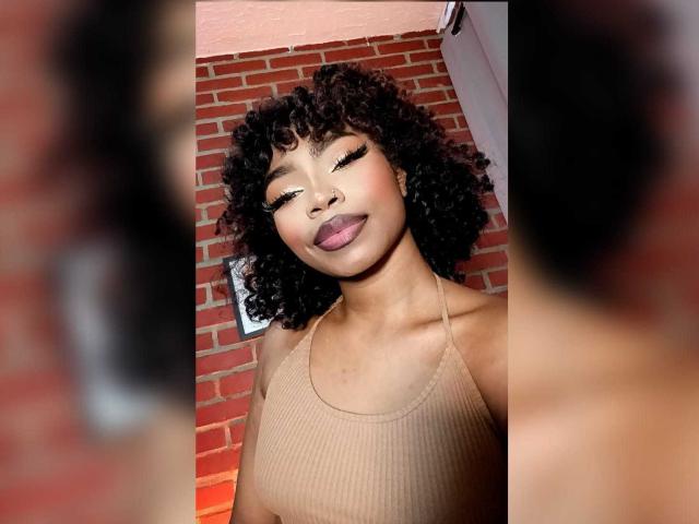 Tanysha69 with curly black hair, tan ribbed top, and purple lipstick, smiling with eyes closed, showing her cute ass in the background. Image is a selfie, face forward.