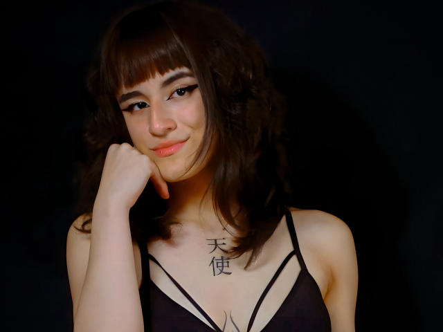 Dalila Cooper, com cabelo castanho ondulado, olhos escuros e um leve sorriso, vestindo uma blusa preta com alças finas, com o queixo apoiado na mão, mostrando uma tatuagem no peito..
