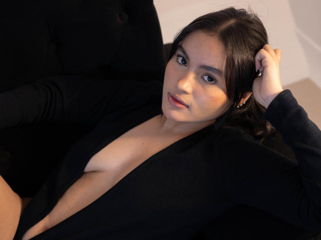 MiiaPalmer with dark hair, wearing a black, deep V-neck, exposing her small, bare tits, lying on a black, tufted couch, looking at the camera with a subtle smile.