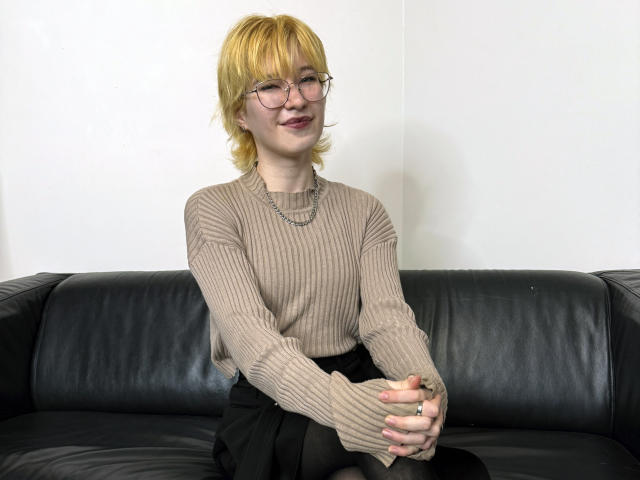 DannaSensual, rubia, con gafas, suéter beige de punto, falda negra, manos juntas, sofá de cuero negro, sonrisa sutil, collar de plata, piel clara, cabello corto, anillo en la mano derecha..