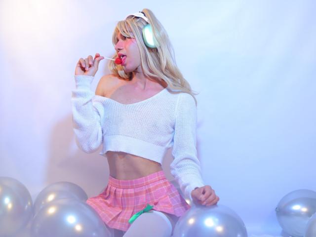 VeronicaBlossom with blonde hair, white off-shoulder sweater, pink plaid skirt, sucking on a red lollipop, surrounded by silver balloons, wearing white headphones.