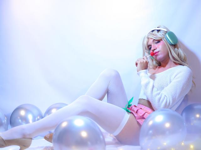 VeronicaBlossom with blonde hair, white sweater, pink shorts, and white thigh-highs, sitting among silver balloons, wearing green headphones, sucking red lollipop, legs crossed.