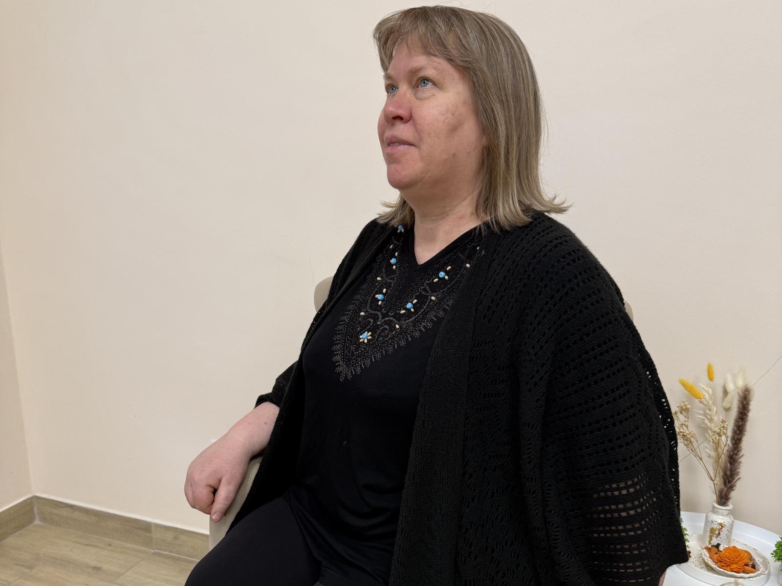 FionaCoode, een vrouw van middelbare leeftijd met kort, grijsbruin haar, gekleed in een zwarte blouse en een zwarte gebreide cardigan, zittend, met haar blik naar de zijkant gericht, draagt een kleine blauw-witte ketting, met een lichte glimlach..