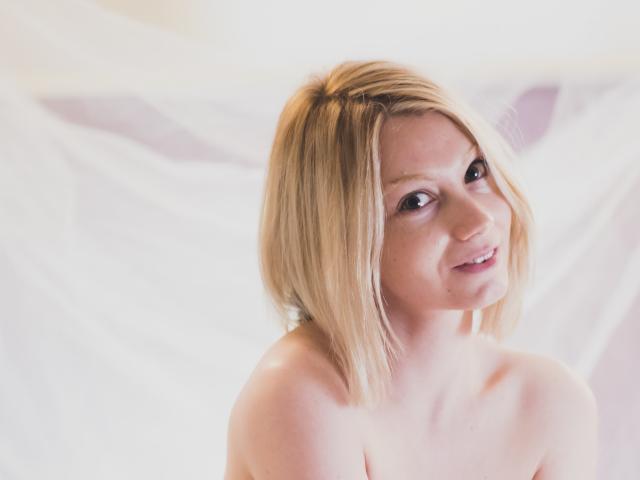 Blonde AfroditaMoore with short hair, smiling, topless, showing bare shoulders, soft light, white background, looking at camera, slight tilt to head, gentle expression.