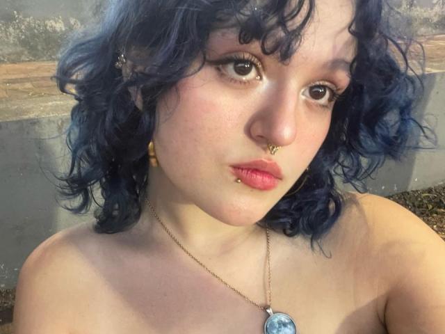 StephyStar with curly blue hair, nose ring, lip piercings, and pink lipstick, wearing a necklace with a blue pendant, has a serious expression and bare shoulders.