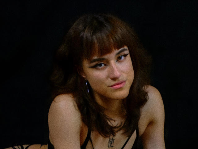 Dalila Cooper, avec ses cheveux bruns foncés ondulés et sa frange, porte du noir, regarde légèrement vers le bas avec un sourire discret, montrant sa poitrine et un petit collier sur un fond noir..