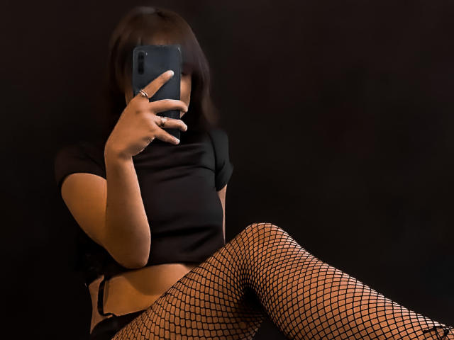 Dalila Cooper, en collants résille, avec un haut noir et un short noir, prend un selfie avec son téléphone, en cachant son visage, tout en montrant son ventre nu et une bague à sa main gauche..