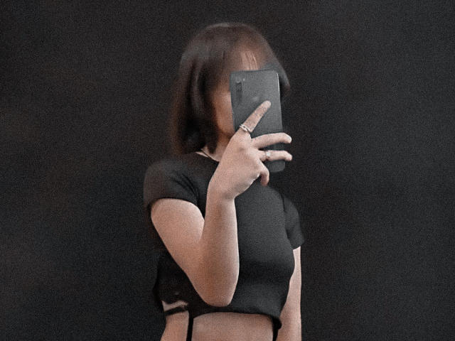 DalilaCooper avec une coupe de cheveux courte brune, un haut court noir, tenant un téléphone devant son visage, un doigt levé, fond noir, une bague visible sur l'index..