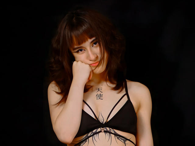 Dalila Cooper mit welligem, braunem Haar, einem schwarzen, trägerlosen BH und einem Tattoo über ihren Brüsten, die ihr Kinn in ihre Hand legt und leicht lächelt. Dunkler Hintergrund..