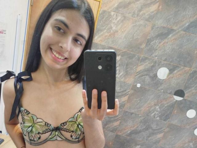 Anna Alvarez mit schwarzen Haaren, lächelnd, hält ein Handy in einem Spiegel-Selfie, trägt einen BH mit Blumenmuster in Grün und Schwarz, wobei ihre kleinen Brüste sichtbar sind..