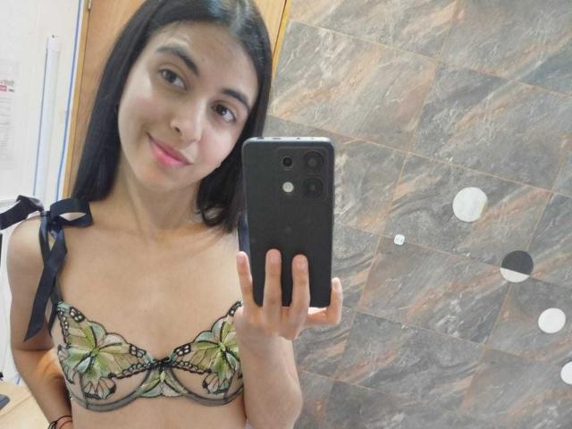 Anna Alvarez tomando un selfie con un teléfono negro, vistiendo un sujetador floral verde y negro, sonriendo, con cabello oscuro y ojos marrones, sosteniendo el teléfono con la mano derecha..