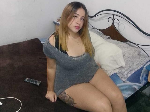 DahianaX met lang bruin haar, gekleed in een grijze jurk met een open schouder, met een zwarte bh en een tatoeage op haar rechter dij, zittend op een bed met een afstandsbediening, een kussen en een bruine deken..