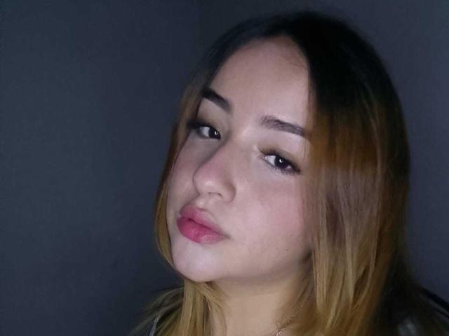 DahianaX a la peau claire, les yeux marron et des lèvres légèrement entrouvertes de couleur rose. Elle regarde sur le côté avec une expression subtile, presque endormie. Ses cheveux blonds sont lisses..
