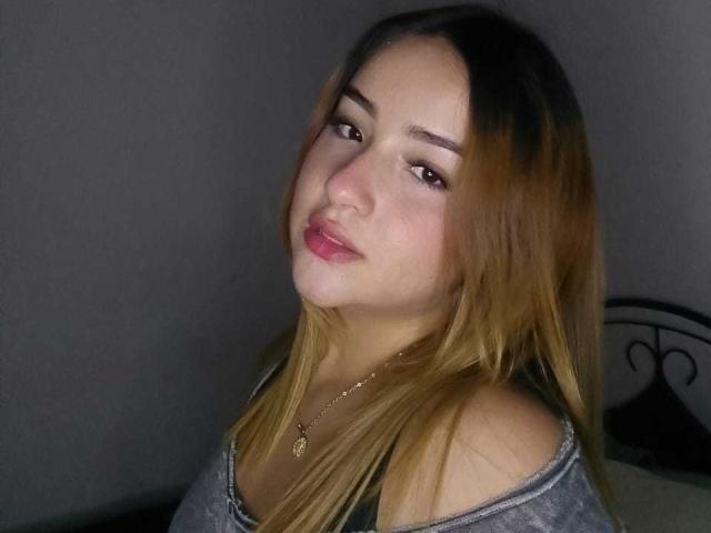DahianaX has light brown hair, a cute face, and is looking slightly to the side with a subtle smile. She's wearing a gray off-shoulder top and a gold necklace.