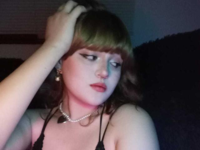 StephyStar with green-brown hair, bangs, and a nose ring, wearing black lace top, pearl necklace, and earrings, hand in hair, looking down with a slightly annoyed expression.