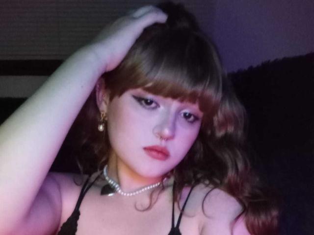 StephyStar with wavy brown hair, black eyeliner, nose ring, pearl necklace, and earrings, hand in hair, looking at camera, in low light, showing tits and a serious face.