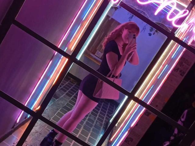StephyStar with pink hair, black short dress, pink purse, and black shoes, taking a mirror selfie, neon lights in background, holding phone, reflection shows legs and neon glow.
