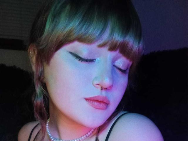 StephyStar has blue-tinted skin, closed eyes, and a nose ring. She's wearing a black strap top with pink lipstick and a pearl necklace. Her brown hair has bangs.