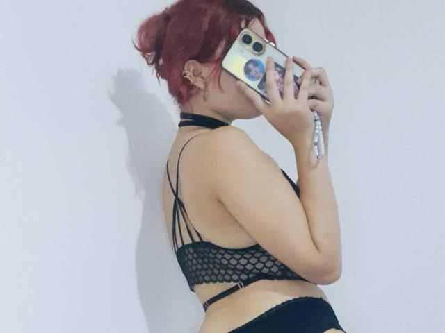 StephyStar with red hair, wearing a black fishnet bra, holding a phone, covering her face, black choker, black pants, white wall, shadow on the wall, holding phone with stickers.