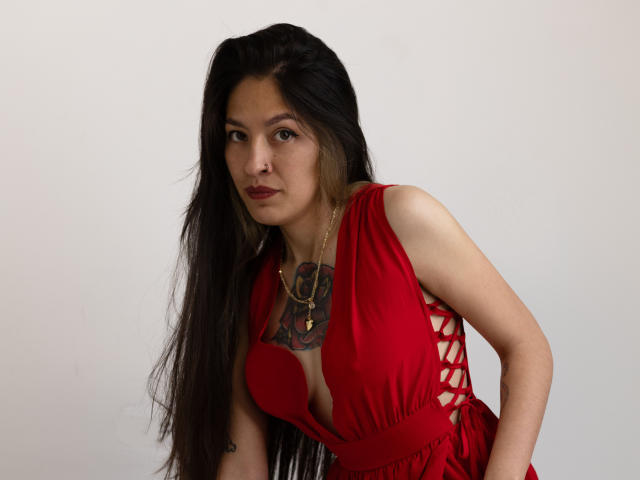 AuroriaSforza, avec ses longs cheveux noirs, sa robe rouge avec des détails en dentelle sur le côté, qui dévoile sa poitrine, et un tatouage de rose sur sa poitrine, portant un collier en or, regardant l'appareil photo..