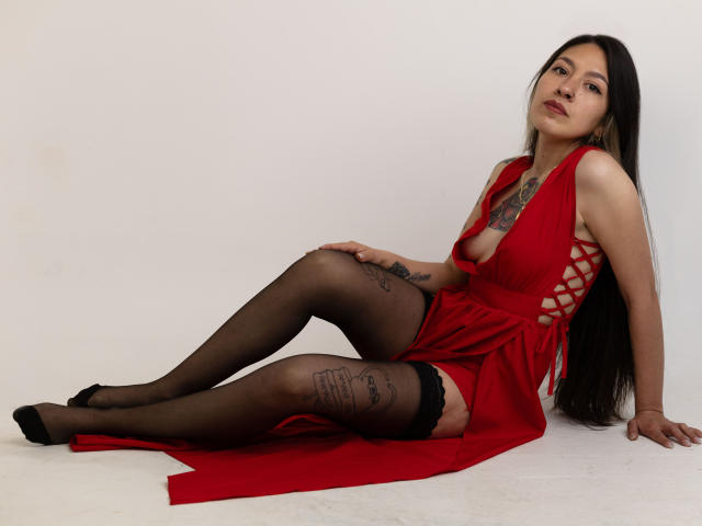 AuroriaSforza indossa un abito rosso con lacci laterali, calze nere e tacchi neri, seduta con le gambe divaricate, mostrando tatuaggi sulle cosce, lunghi capelli neri e un'espressione sicura..