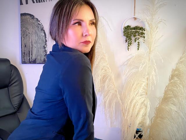 AdelaShery, avec ses cheveux bruns mi-longs, portant un costume bleu, regardant par-dessus son épaule avec une expression sérieuse, devant de l'herbe blanche et duveteuse et une plante suspendue..