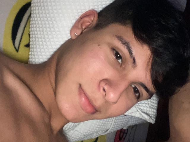 WillStarr's face, shirtless, with dark hair, brown eyes, nose piercing, and ear stud, lying on a white pillow, showing a slight smile. Soft, even skin, relaxed.