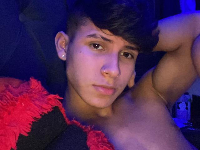 WillStarr with short black hair, brown eyes, nose ring, and ear piercing, shirtless, lying down, arm raised, looking at camera, red and black fuzzy blanket in the foreground.