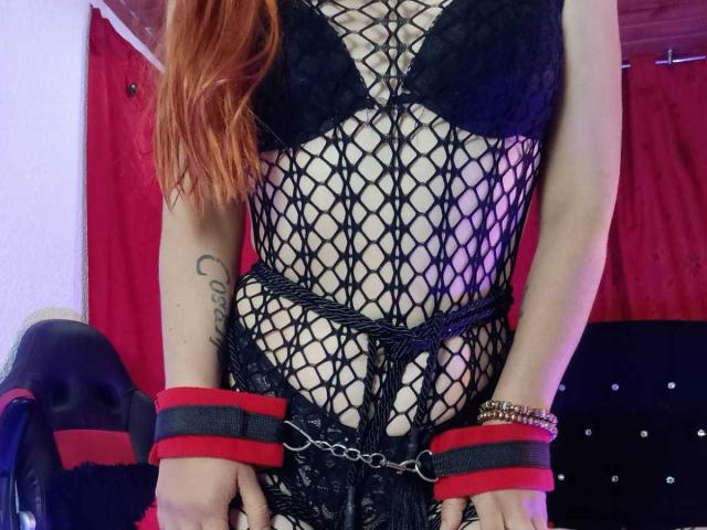 SelenneMillers with red hair, black fishnet lingerie, black bra, red and black wrist cuffs, tattoo on right arm, black lace, and chain belt. Visible pussy, black lace panties.