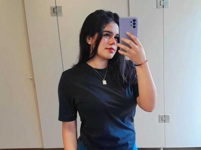 VictoriaBoston, com cabelos longos e pretos, camiseta preta, seios pequenos, segurando um telefone roxo, tirando uma selfie no espelho, usando um colar, olhando ligeiramente para o lado, sorrindo suavemente..