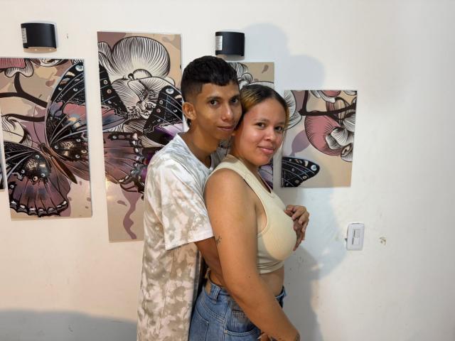 JoshNevalov with short dark hair, wearing a white floral shirt, stands behind JoshNevalov with blonde hair, wearing a beige crop top and blue jeans, embracing each other. Butterfly art in background.
