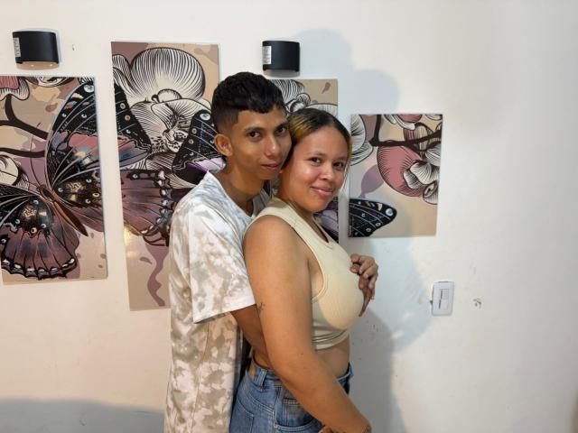 JoshNevalov with short black hair and camo shirt, arms around JoshNevalov with blonde hair, tan skin, white crop top, showing boobs, smiling. Butterfly art in background.