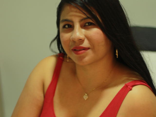 AlissonMyst, avec de longs cheveux noirs, un débardeur rouge, de petites boucles d'oreilles dorées et un collier en forme de cœur, souriant légèrement, montrant sa poitrine de taille moyenne, et portant du rouge à lèvres rose..