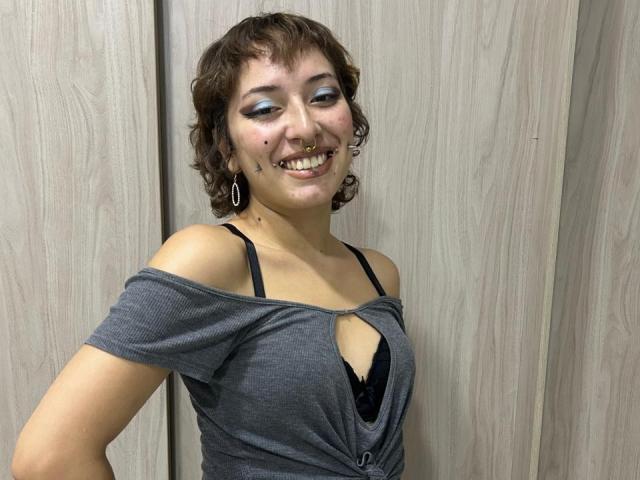 Pasiflor, avec ses cheveux bruns bouclés, son fard à paupières bleu et son anneau de nez, souriante, portant un haut gris à une épaule, laissant apparaître un soutien-gorge noir et sa poitrine, debout devant des panneaux en bois..