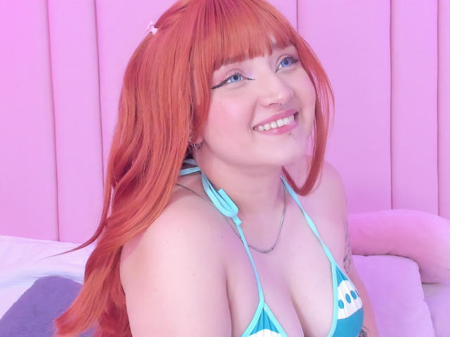 Tracie Lowe, avec ses cheveux roux, souriante, portant un haut de maillot de bain bleu, montrant ses seins, avec un eye-liner bleu, un anneau au nez et un joli sourire, assise sur un lit avec un éclairage rose..