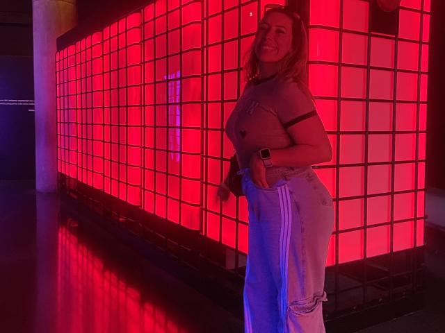 JennyLawry with a cute ass, wearing a grey shirt and ripped blue jeans, standing in front of a red illuminated grid wall, smiling at the camera.