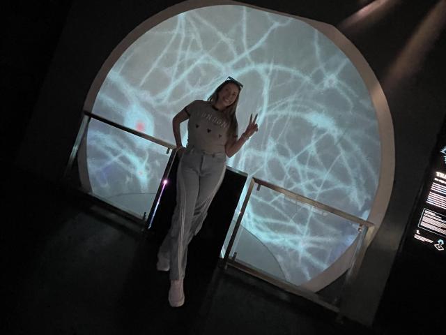 JennyLawry se tient debout, souriante, vêtue d'un t-shirt gris, d'un pantalon blanc et de baskets roses, et fait un signe de la paix. Une projection lumineuse avec des motifs blancs en forme de toile d'araignée se trouve derrière elle..