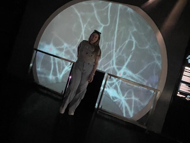 JennyLawry stands in a dark room, wearing a beige military-style dress with buttons, white boots, and a cap, against a white screen with cracked light patterns.