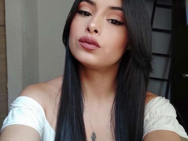 EmilyVv has long black hair, pouty pink lips, and a white off-shoulder top, showing a subtle tattoo near her chest, with a slight, sexy, sultry expression.
