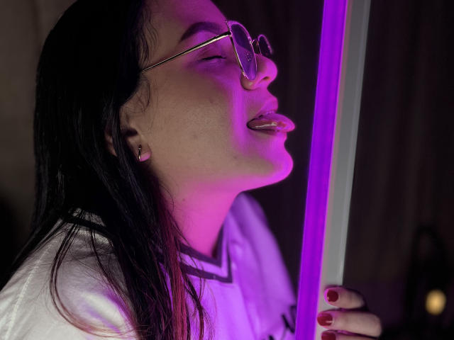 Minddy with black hair, glasses, and tongue out, holding a purple neon rod, wearing a white shirt, pinkish-purple light on her face, red nail polish.
